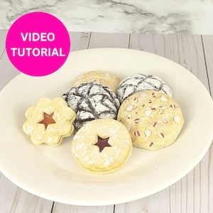 May include: A white ceramic plate holds an assortment of decorated cookies. The cookies vary in shape and design, including star-shaped cutouts and powdered sugar dusting. A pink circle with the words "VIDEO TUTORIAL" is in the upper left corner.