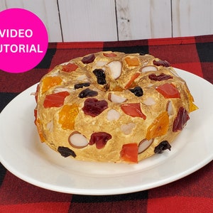 Peut inclure: Un gâteau aux fruits en forme d'anneau avec une variété de fruits secs et de noix, notamment des cerises, des abricots et des amandes. Le gâteau est brun doré et a une finition légèrement glacée.