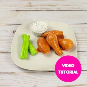 Puede incluir: Un plato blanco con cinco alitas de pollo doradas, dos tallos de apio verdes, un pequeño tazón blanco con salsa blanca y el texto "VIDEO TUTORIAL" en rosa sobre un círculo rosa.