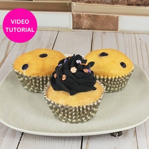 May include: Three cupcakes on a white plate. Two cupcakes have yellow frosting with dark spots. The center cupcake has black frosting with decorative sprinkles. The cupcakes are in patterned paper liners. A pink circle with the words "VIDEO TUTORIAL" is in the upper left.