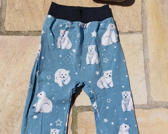 Op de afbeelding: Babybroek met een lichtblauwe achtergrond en een patroon van witte ijsberen en witte sterren. De broek heeft een zwarte tailleband en zwarte manchetten. Een beschilderde steen met het woord "handgemacht" staat op de achtergrond.