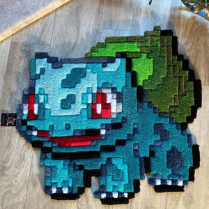 Può includere: Un tappeto pixelato raffigurante il Pokémon Bulbasaur, nei toni del turchese, verde, rosso e nero. Il tappeto ha un aspetto strutturato ed è posato su un pavimento in legno. Il design ricorda la grafica dei videogiochi classici.