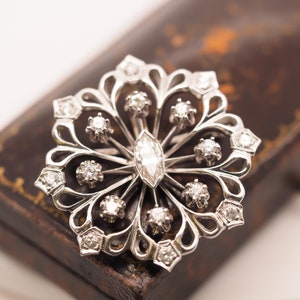 May include: A silver brooch with a floral design and multiple diamonds. The brooch is sitting on a brown leather box.
