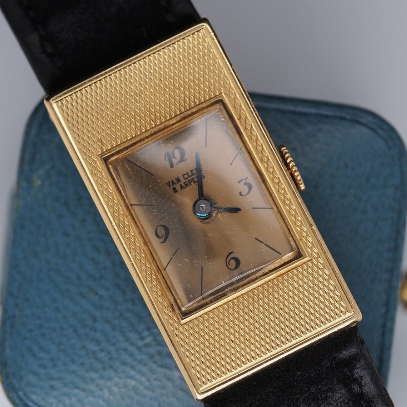 May include: A gold-tone rectangular wristwatch with a black leather band. The watch face is white with black Roman numeral hour markers and black hands. The watch face also has the text "Van Cleef & Arpels" printed on it.