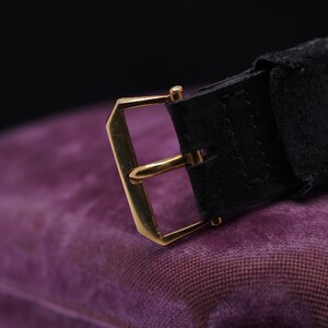 May include: A gold-toned watch buckle with a black leather strap. The buckle is rectangular with a smooth, polished finish. The strap is made of a soft, supple leather.