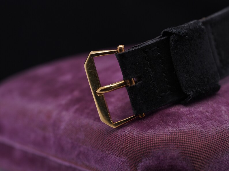May include: A gold-toned watch buckle with a black leather strap. The buckle is rectangular with a smooth, polished finish. The strap is made of a soft, supple leather.