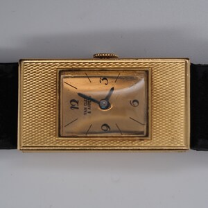 May include: A gold-toned wristwatch with a square face and a black leather band. The watch face has Roman numeral markings and the text "Van Cleef & Arpels" is visible.