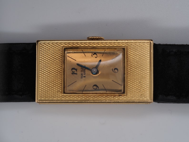 May include: A gold-toned wristwatch with a square face and a black leather band. The watch face has Roman numeral markings and the text "Van Cleef & Arpels" is visible.
