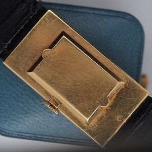 May include: A gold-toned watch buckle with a black leather strap. The buckle has a rectangular shape with a raised, rectangular section in the center. The buckle is engraved with the number "1820".