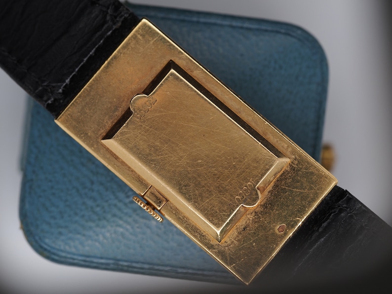 May include: A gold-toned watch buckle with a black leather strap. The buckle has a rectangular shape with a raised, rectangular section in the center. The buckle is engraved with the number "1820".