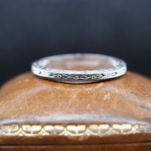 Puede incluir: Una fina alianza de plata con un diseño floral intrincado. La alianza está sobre una caja de anillo de cuero marrón con un borde dorado.