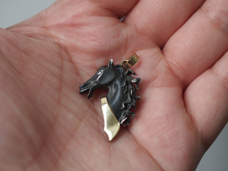K&ouml;nnte beinhalten: Ein silberner Pferdekopf-Anh&auml;nger mit einem goldenen Akzent am Hals. Der Anh&auml;nger ist detailliert mit einer strukturierten M&auml;hne und einem realistischen Pferdekopf.