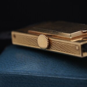 May include: A gold-toned wristwatch with a rectangular case and a textured band. The watch has a small, round crown on the side.