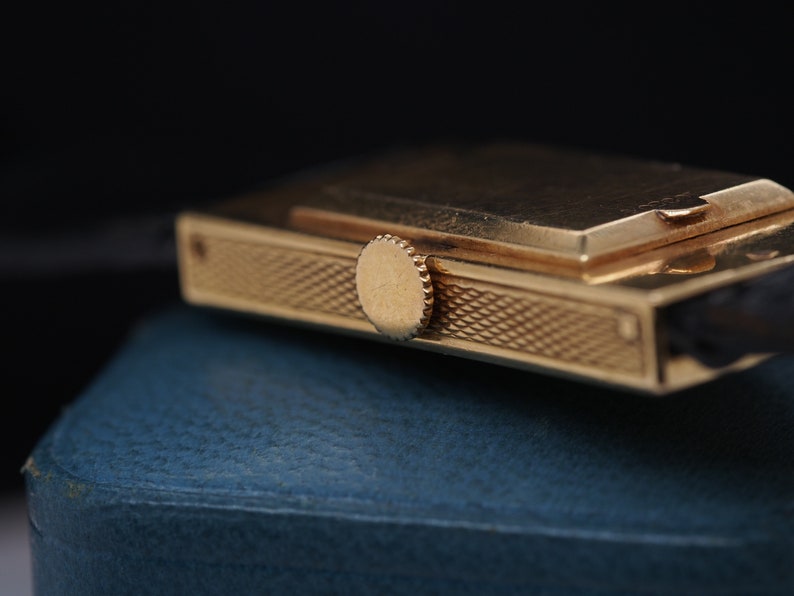 May include: A gold-toned wristwatch with a rectangular case and a textured band. The watch has a small, round crown on the side.