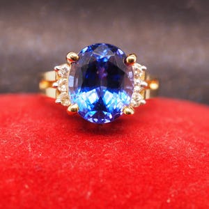 May include: A gold ring featuring a large, oval-cut, deep blue gemstone set in the center. The central stone is flanked on each side by three small, clear, round-cut accent stones. The band is made of polished yellow gold with a classic prong setting.