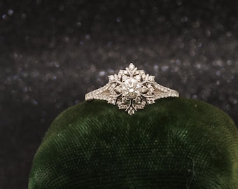 Può includere: Un anello color argento con una pietra centrale a taglio rotondo circondata da un gruppo di pietre più piccole incastonate a pavé disposte in un motivo simmetrico a fiocco di neve, con pietre aggiuntive lungo la fascia divisa.