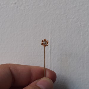 Puede incluir: Un alfiler dorado con un peque&ntilde;o strass transparente engastado en un dise&ntilde;o con forma de flor.