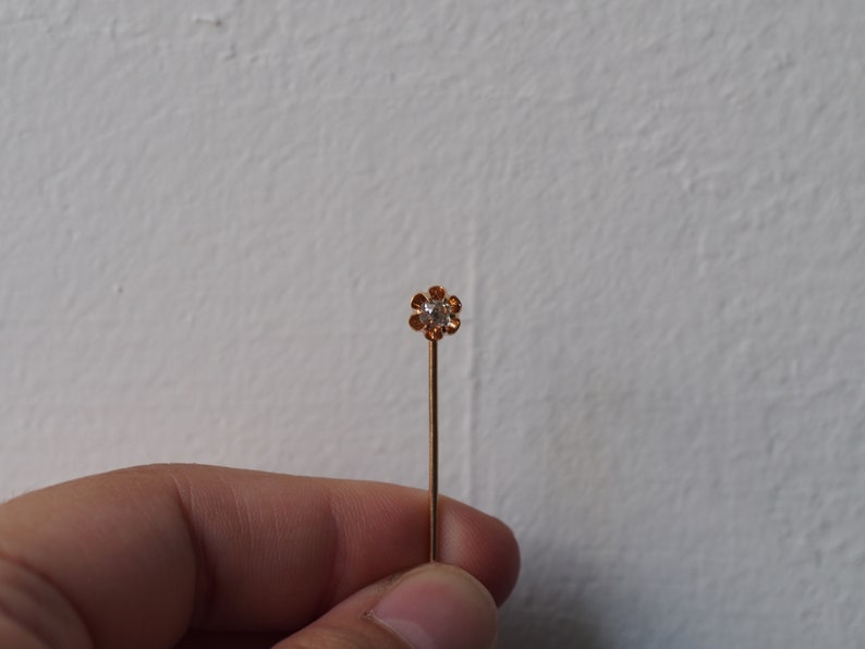 Puede incluir: Un alfiler dorado con un peque&ntilde;o strass transparente engastado en un dise&ntilde;o con forma de flor.