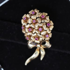Può includere: Spilla floreale dorata con un gruppo di fiori con piccole gemme rosa rotonde al centro. La spilla include un dettaglio a fiocco alla base. Il gioiello è posto su uno sfondo nero.