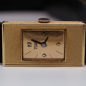 May include: A gold-toned wristwatch with a square face and black hands. The watch face has black Roman numeral hour markers and the text "Van Cleef & Arpels" printed on it.