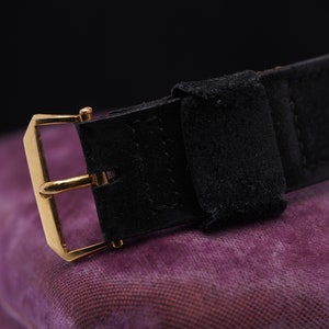 May include: A black leather watch strap with a gold buckle. The strap is on a purple fabric surface.