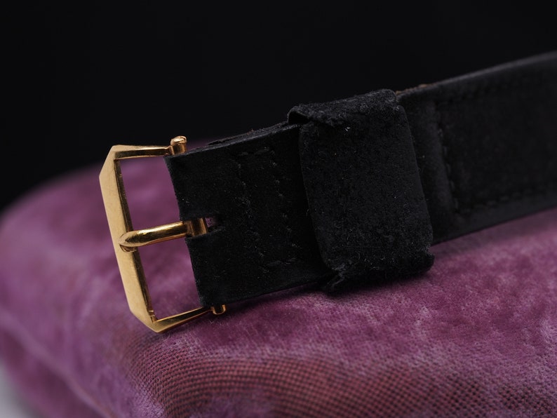 May include: A black leather watch strap with a gold buckle. The strap is on a purple fabric surface.