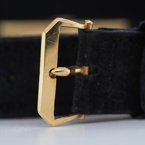May include: A gold-toned watch strap buckle with a black leather strap. The buckle has a simple, rectangular design with a smooth finish.