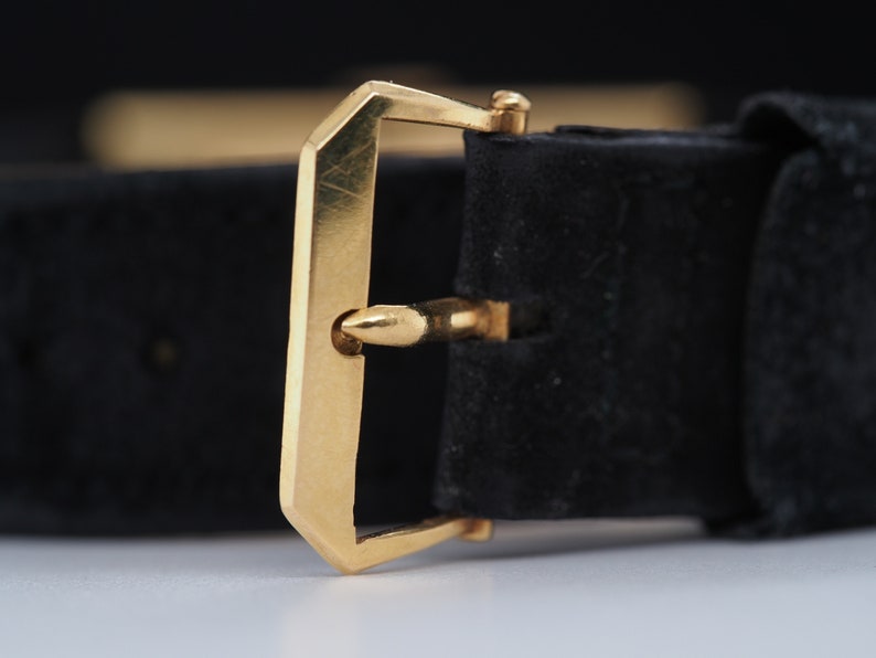 May include: A gold-toned watch strap buckle with a black leather strap. The buckle has a simple, rectangular design with a smooth finish.