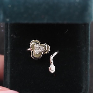 May include: A silver ring with a floral design, featuring three small, clear stones at the center of each petal. The ring is displayed in a dark green velvet jewelry box, with a matching silver earring.