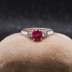 May include: A silver-toned ring featuring a central oval-cut deep red ruby gemstone held in a four-prong setting, flanked by two smaller round-cut clear white diamonds on each side, set on a polished metallic band.