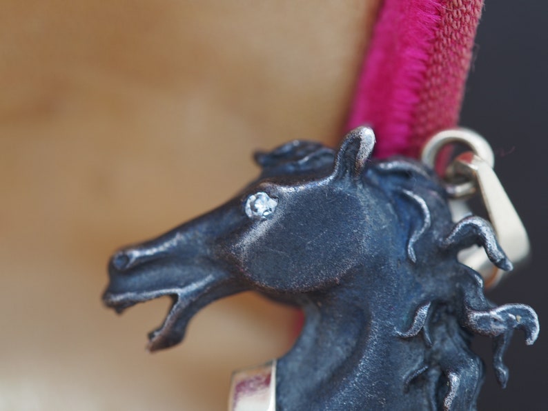 K&ouml;nnte beinhalten: Ein silberner Pferdekopf-Anh&auml;nger mit einem einzigen klaren Strassstein-Auge. Die M&auml;hne des Pferdes ist detailliert und der Anh&auml;nger ist an einer silbernen Kette befestigt.