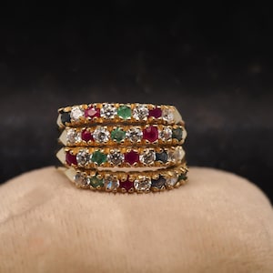 May include: A stack of four gold rings, each featuring a row of alternating round-cut gemstones including clear white stones, red rubies, green emeralds, and dark blue sapphires set in a continuous band design across the front of each ring.