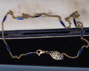 Puede incluir: Un collar dorado con detalles de esmalte azul. El collar presenta segmentos ovalados de esmalte azul unidos por una delicada cadena. El cierre incluye un elemento decorativo con un centro de esmalte azul. El collar se muestra en una caja azul.