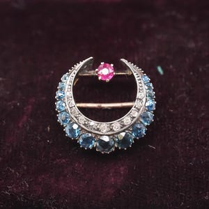 Puede incluir: Un broche de plata en forma de media luna con una piedra roja en el centro y piedras azules alrededor del borde.