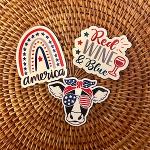 May include: Three patriotic themed stickers. One sticker features a rainbow with stars and the text "america". Another sticker features a cow with sunglasses and a bandana. The third sticker features a wine glass and the text "Red Wine & Blue".