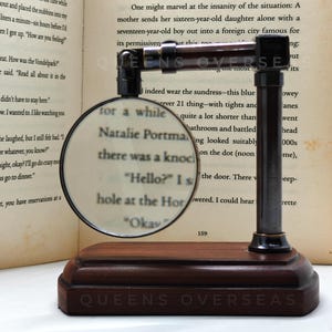 May include: A magnifying glass with a dark wooden base and metal frame, resting on a book. The magnifying glass is focused on text, and the base has the text "QUEENS OVERSEAS".