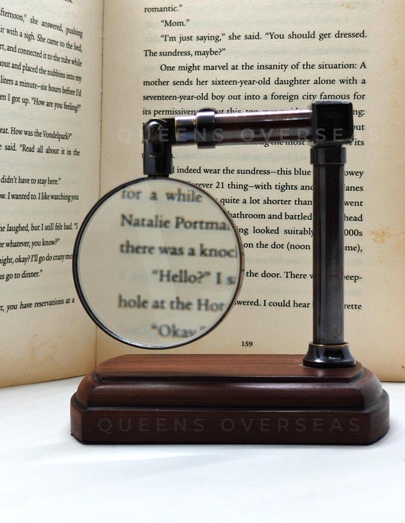 May include: A magnifying glass with a dark wooden base and metal frame, resting on a book. The magnifying glass is focused on text, and the base has the text "QUEENS OVERSEAS".