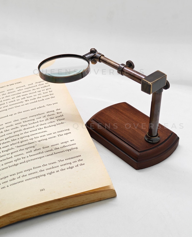 May include: A vintage-style magnifying glass with a dark wooden base and a bronze-colored adjustable arm. The magnifying glass is positioned over an open book, ideal for reading or examining small details. The base measures approximately 6 inches in length.