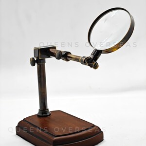 May include: A vintage-style magnifying glass with a dark brown wooden base and a brass-colored frame. The magnifying glass is mounted on an adjustable arm. The text "QUEENS OVERSEAS" is visible on the base.