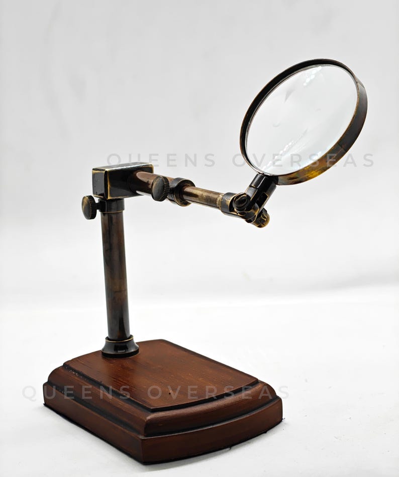 May include: A vintage-style magnifying glass with a dark brown wooden base and a brass-colored frame. The magnifying glass is mounted on an adjustable arm. The text "QUEENS OVERSEAS" is visible on the base.