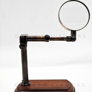 May include: A vintage-style magnifying glass with a dark wooden base and a brass-colored frame. The magnifying glass is round and mounted on an adjustable arm. The base is rectangular and has a dark brown finish.