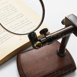 May include: A vintage-style magnifying glass with a dark brown wooden base and a brass-colored frame. The magnifying glass is positioned over an open book. The base has the text "QUEENS OVERSEA".