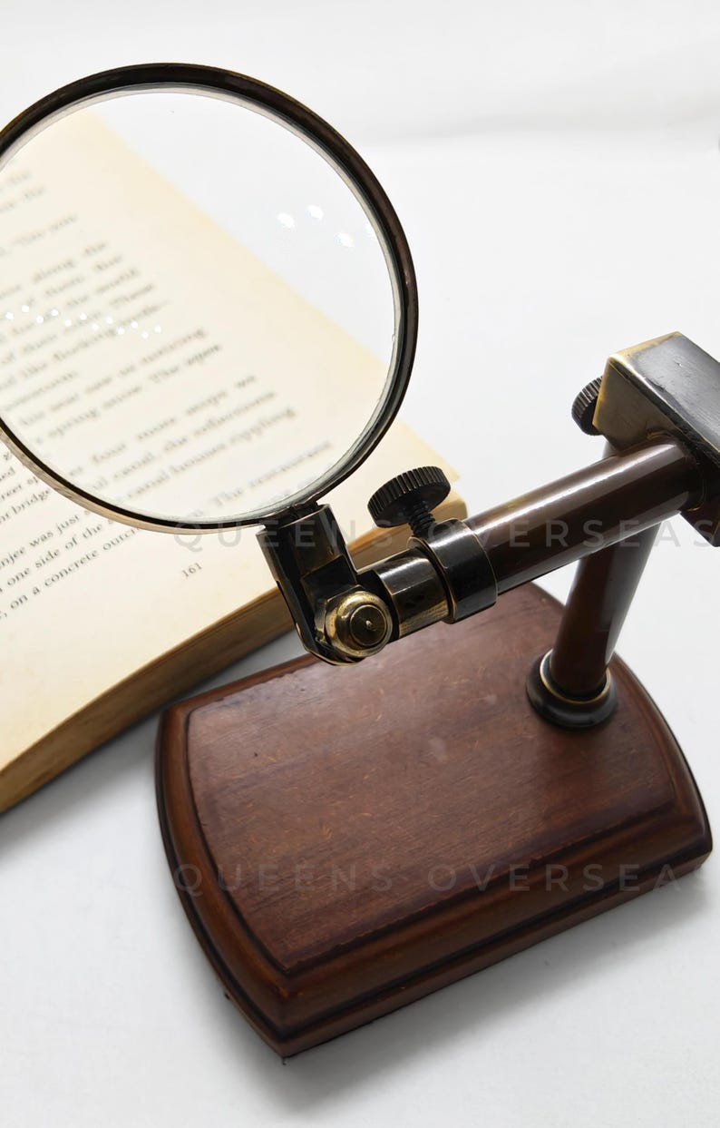 May include: A vintage-style magnifying glass with a dark brown wooden base and a brass-colored frame. The magnifying glass is positioned over an open book. The base has the text "QUEENS OVERSEA".