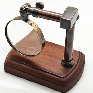 May include: A vintage-style magnifying glass with a wooden base and a brass frame. The magnifying glass is mounted on a dark brown metal arm and is adjustable. The base has the text "QUEENS OVERSEAS".