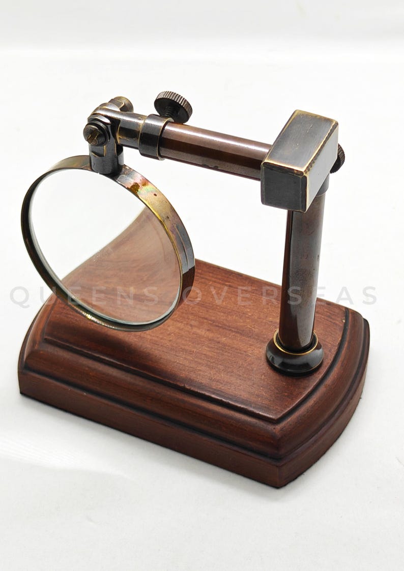 May include: A vintage-style magnifying glass with a wooden base and a brass frame. The magnifying glass is mounted on a dark brown metal arm and is adjustable. The base has the text "QUEENS OVERSEAS".