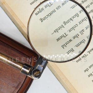 May include: A vintage-style magnifying glass with a brass handle and a wooden base, resting on an open book. The magnifying glass is focused on the text, enhancing the visibility of the words. The text in the image reads "The iepen".