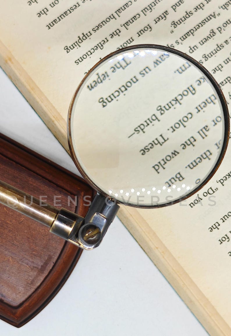 May include: A vintage-style magnifying glass with a brass handle and a wooden base, resting on an open book. The magnifying glass is focused on the text, enhancing the visibility of the words. The text in the image reads "The iepen".