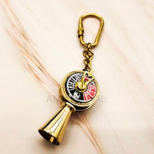 Può includere: Portachiavi a forma di ruota di nave in ottone con quadrante rosso e nero e un fischietto in ottone. Il portachiavi ha una catena dorata e un anello diviso.