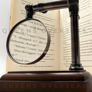 May include: A vintage-style magnifying glass with a dark brown wooden base and frame, resting on an open book. The magnifying glass is round with a dark metal rim, and the text "QUEENS OVERSEAS" is visible on the base.