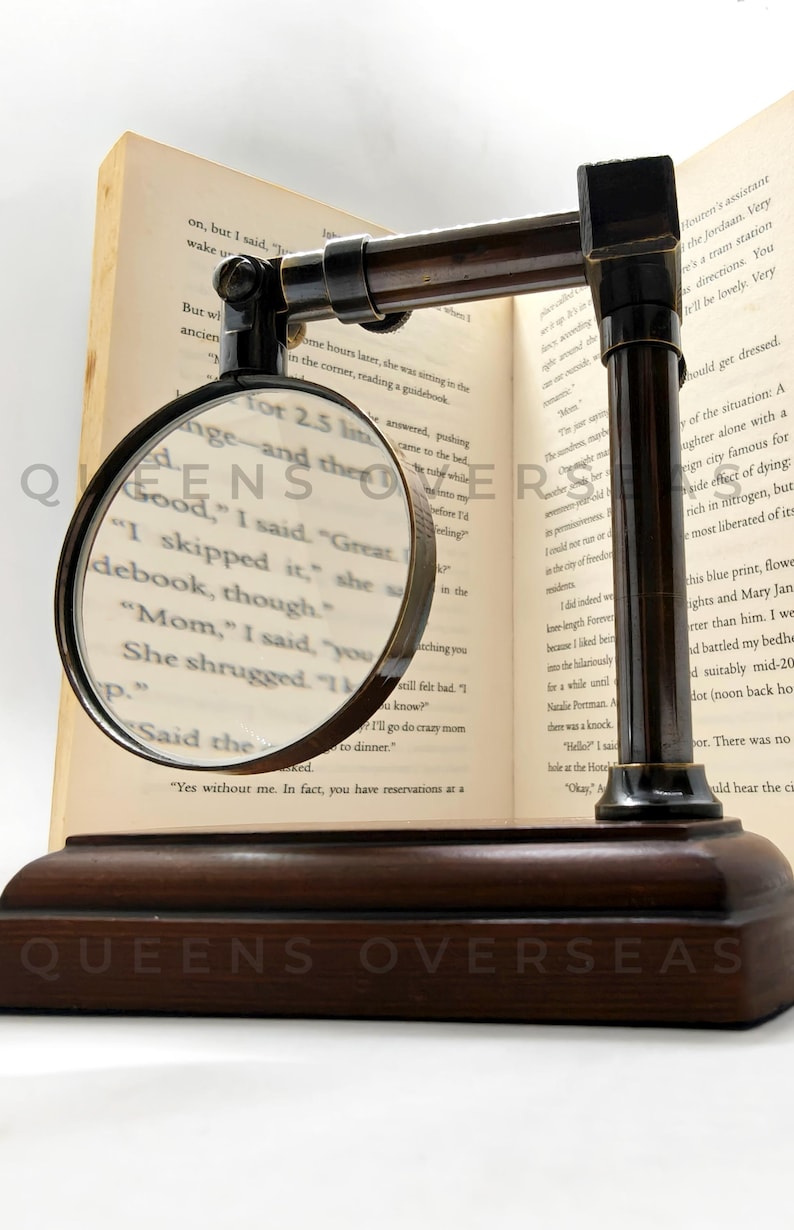 May include: A vintage-style magnifying glass with a dark brown wooden base and frame, resting on an open book. The magnifying glass is round with a dark metal rim, and the text "QUEENS OVERSEAS" is visible on the base.